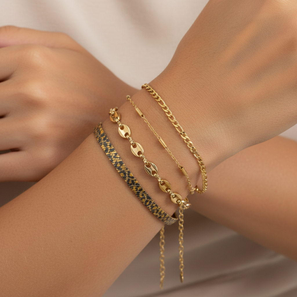Four gold-tone Ready-to-Wear (RTW) bracelets layered on a model's wrist, including a snake chain, a coffee bean chain, a delicate bar chain, and a curb chain. Focus on the jewellery pieces.